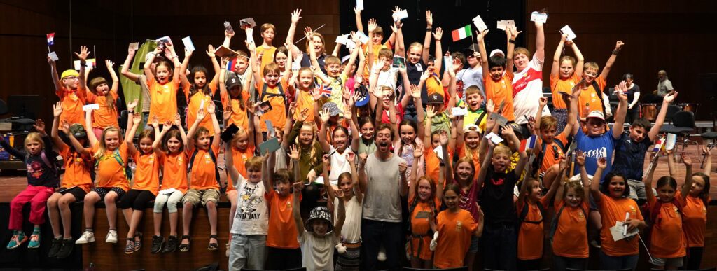 Bunte Gruppe von Kindern in orangefarbenen T-Shirts, die begeistert auf der Bühne der Stadtkapelle Wernau posieren und dabei die Arme heben. Die Szene vermittelt Freude an der Musik und spricht Musiker sowie Musikliebhaber im Raum Esslingen und Stuttgart an. Ideal für Suchanfragen zu musikalischen Themen und Veranstaltungen im Landkreis Esslingen und Stuttgart.