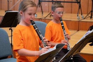 Zwei Kinder in orangefarbenen T-Shirts spielen Klarinette während einer Probe der Stadtkapelle Wernau. Die Szene zeigt die Förderung der Musikausbildung für junge Talente im Raum Esslingen und Stuttgart.