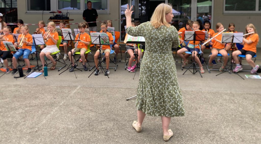Die Leiterin der Stadtkapelle Wernau dirigiert eine Gruppe junger Musiker in leuchtend orangefarbener Kleidung während einer Aufführung im Freien. Die Veranstaltung richtet sich an Musikliebhaber im Raum Esslingen und Stuttgart und fördert das musikalische Engagement der Kinder.