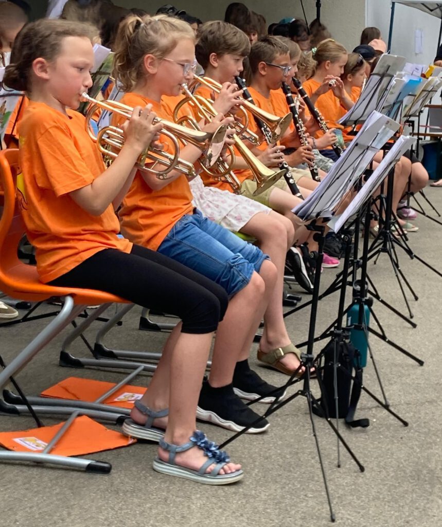 Musiker der Stadtkapelle Wernau spielen bei einem Auftritt im Freien, gekleidet in orangefarbene T-Shirts. Die Gruppe besteht hauptsächlich aus Kindern und Jugendlichen, die verschiedene Blasinstrumente wie Trompeten und Klarinetten spielen. Ideal für Musikliebhaber in Esslingen und Stuttgart, um mehr über lokale musikalische Veranstaltungen und Talente zu erfahren.