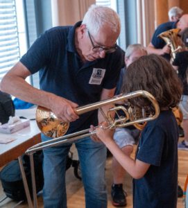 Ein erfahrener Musiker der Stadtkapelle Wernau zeigt einem Kind das Spielen der Posaune während eines Musikworkshops. Die Veranstaltung richtet sich an Musiker und Musikliebhaber im Raum Esslingen und Stuttgart und fördert die musikalische Bildung in der Region.