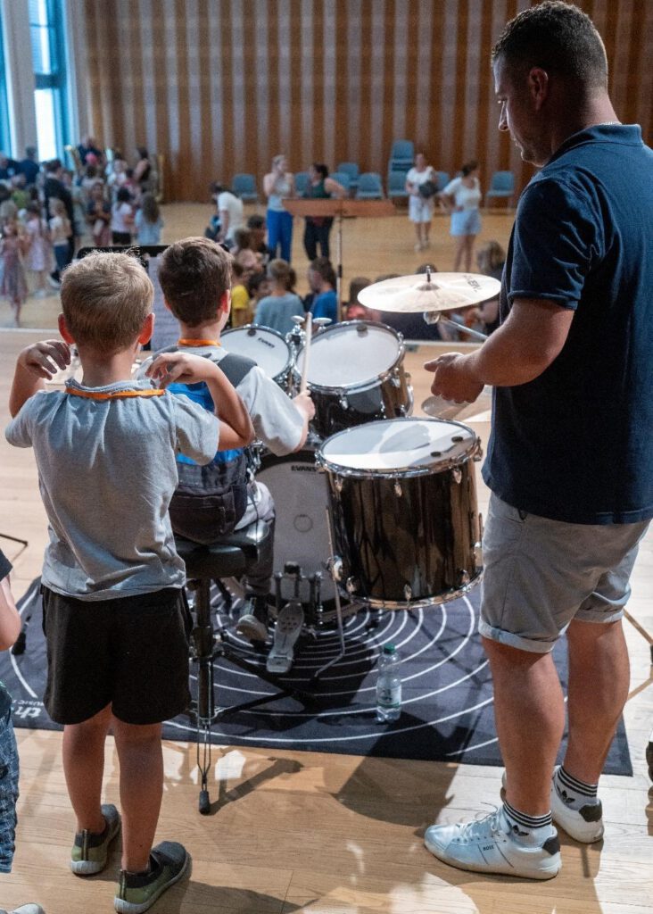 Auf dem Bild sind Kinder zu sehen, die auf einer Bühne zusammen mit einem Erwachsenen an einem Schlagzeug stehen. Die Szene findet während eines musikalischen Events der Stadtkapelle Wernau statt, das Familien und Musikliebhaber im Landkreis Esslingen und Stuttgart anspricht.