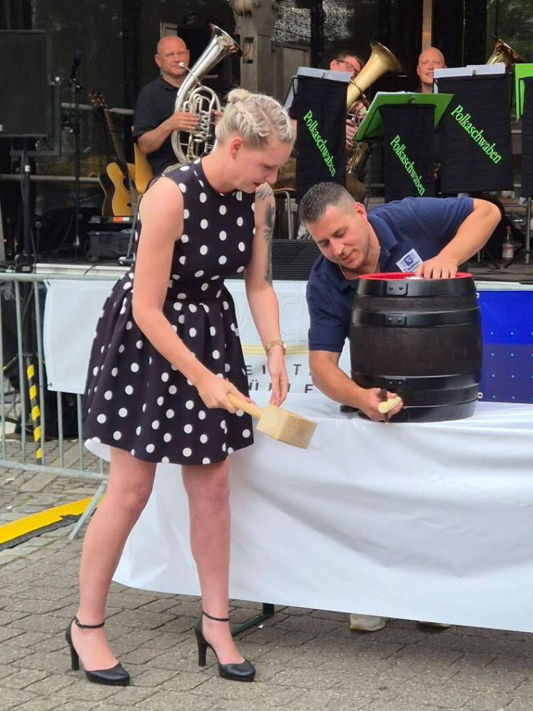Ein Junge und eine Frau klopfen gemeinsam auf ein Holzfass während eines musikalischen Events der Stadtkapelle Wernau. Im Hintergrund spielen Musiker auf verschiedenen Instrumenten. Die Szene zeigt eine lebendige Atmosphäre und spricht Musikliebhaber im Raum Esslingen und Stuttgart an.