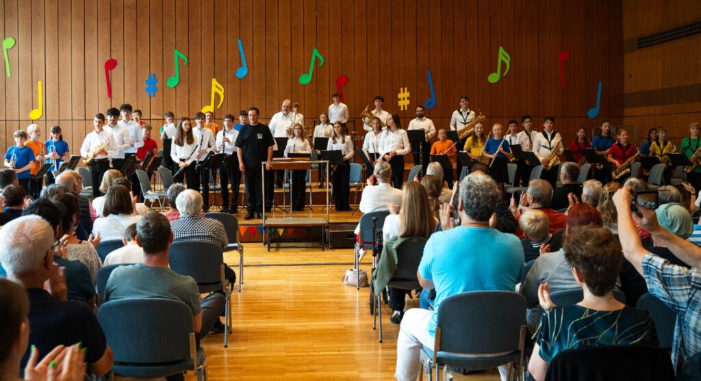 Auftritt der Stadtkapelle Wernau mit jungen Musikern in bunten Outfits während eines Konzerts. Publikum applaudiert im Hintergrund, umrahmt von bunten Notenmotiven an der Wand. Veranstaltung zielt darauf ab, Musiker und Musikliebhaber im Raum Esslingen und Stuttgart anzusprechen.