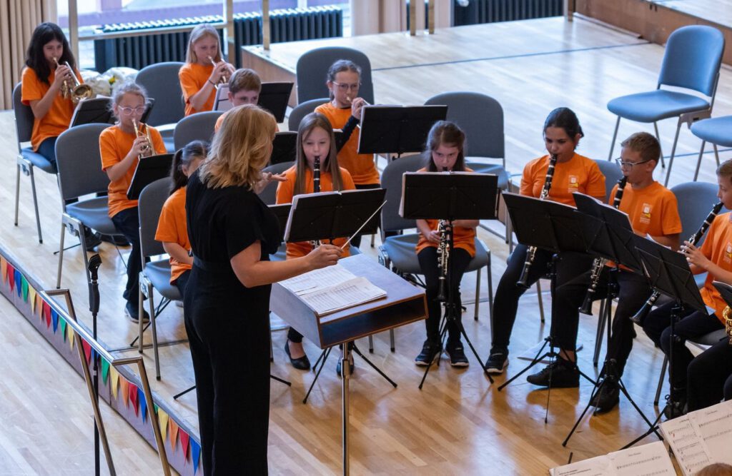 Die Stadtkapelle Wernau probt mit jungen Musikern in orangefarbenen T-Shirts, während eine Dirigentin ihnen Anweisungen gibt. Im Hintergrund sind Notenständer und weitere Musikinstrumente zu sehen. Diese Bilder sollen Musiker und Musikliebhaber im Landkreis Esslingen und Stuttgart ansprechen und auf musikalische Aktivitäten aufmerksam machen.