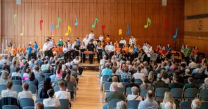 Auftritt der Stadtkapelle Wernau mit jungen Musikern in einem vollen Saal, bunte Musiknoten im Hintergrund, um Musiker und Musikliebhaber aus Esslingen und Stuttgart anzuziehen.