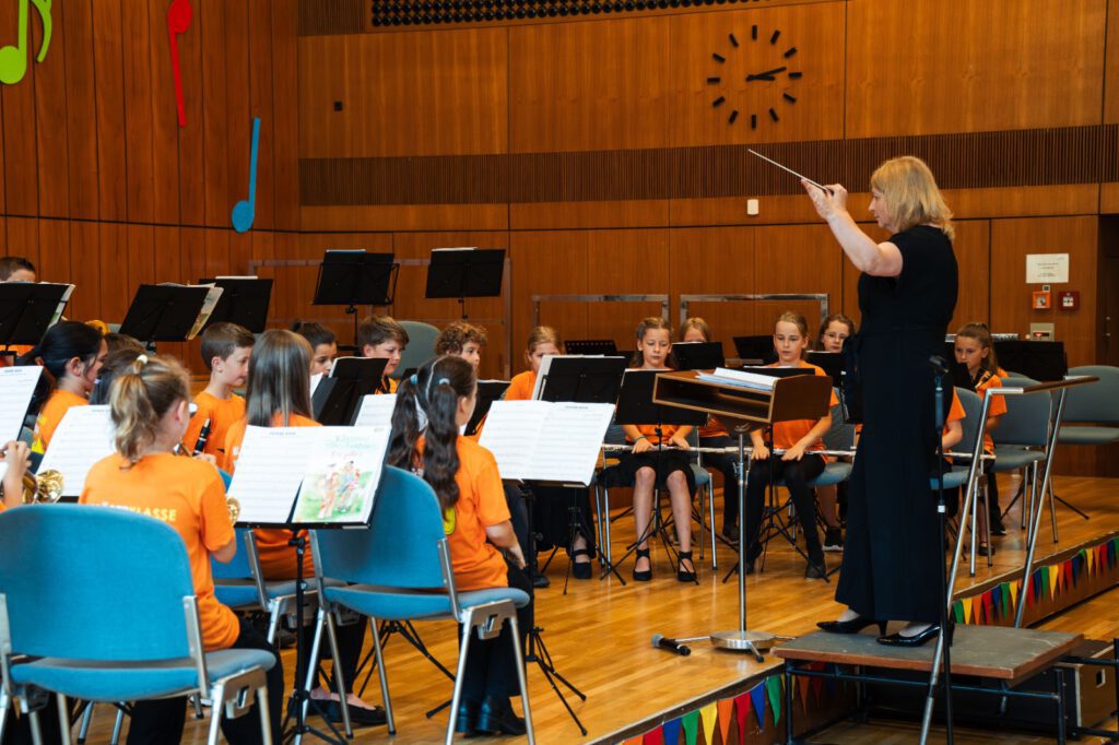 Dirigentin leitet die Stadtkapelle Wernau während einer Probe mit jungen Musikern in orangefarbener Kleidung. Die Bühne ist dekoriert und zeigt Noten sowie musikalische Elemente. Diese Darstellung spricht Musikliebhaber im Landkreis Esslingen und Stuttgart an und fördern das Interesse an musikalischen Aktivitäten und Veranstaltungen.
