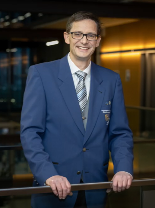 Portrait des Dirigenten der Stadtkapelle Wernau, der Musiker und Musikliebhaber im Raum Esslingen und Stuttgart anspricht. Das Bild zeigt ihn in einem blauen Anzug, lächelnd und stehend in einem modernen Raum, ideal für die Förderung musikalischer Themen.